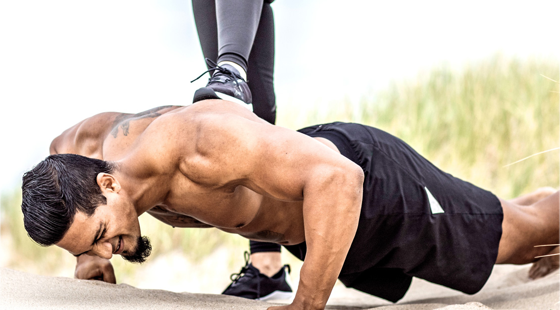 Muscular man performing pushup on the beach while foot is on his back